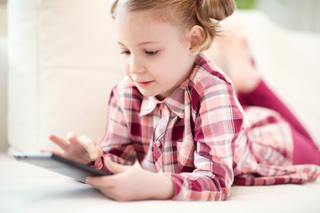 Pretty little child girl using a digital tablet, looking and smiling while lying on the white couch