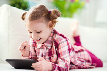Pretty little child girl  using a digital tablet, looking and smiling while lying on the whitw couch