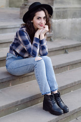 Outdoor portrait of young attractive lady with curly brunette hair in early spring. Model wearing a checkered shirt, jeans and hat, sitting on the stairs and smiling.