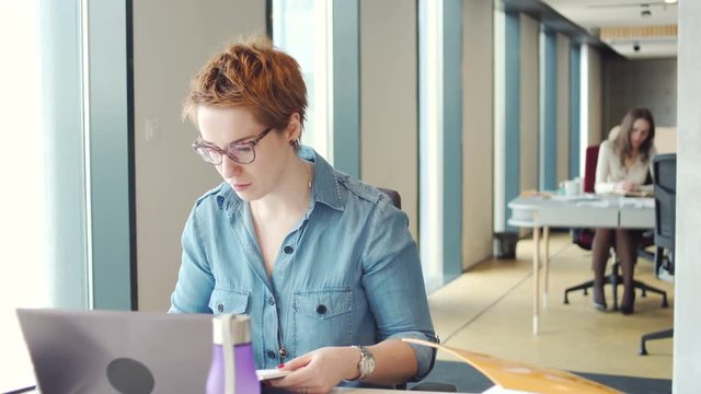young busy woman with short red hair and glasses work diligently on laptop computer with a lot of papers and documents, making notes sitting at the table in co working office with big windows