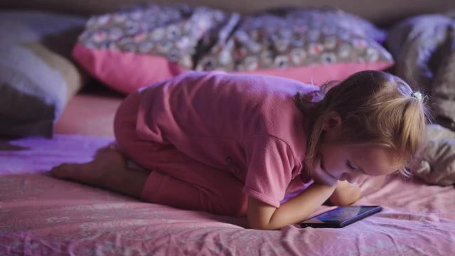 Little girl dressed in pink pyjamas with mobile phone in her hands is lying in bed and watching cartoons. Small toddler female kid is relaxing and playing with cellphone in her bedtime.