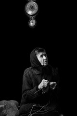 Old woman wearing headscarf, holding yarn strand and knitting needles and looking aside. Black-and-white low key photograph on black background. Blurry and partially visible old clock above her head.