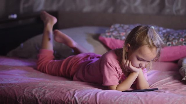 Cute blonde small girl dressed in pink pyjamas is lying in bed and watching cartoons with smartphone. Toddler female kid is relaxing and playing with mobile phone during her bedtime.