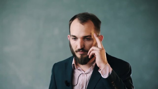 Young caucasian bearded man in blue business jacket and striped shirt with lavalier microphone on isolated background wall shake head, put fingers on temple and talks weary