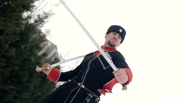 A young man in Cossack clothes swinging a sword in a winter landscape in the snow.
