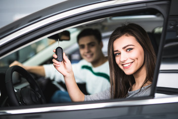 Beautiful young couple sitting at the front seats of their new car while woman showing keys and smiling