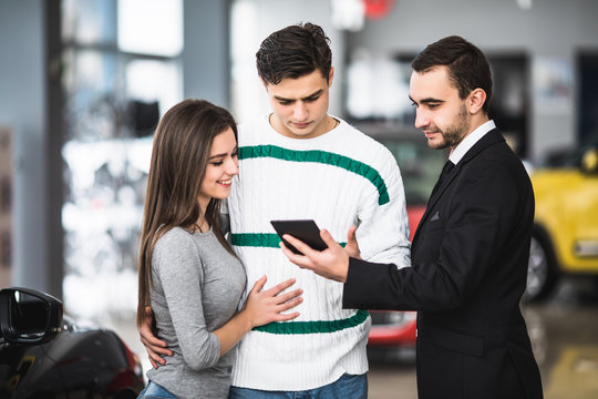 Happy Couple With Car Dealer Look At Tablet In Auto Show Or Salon