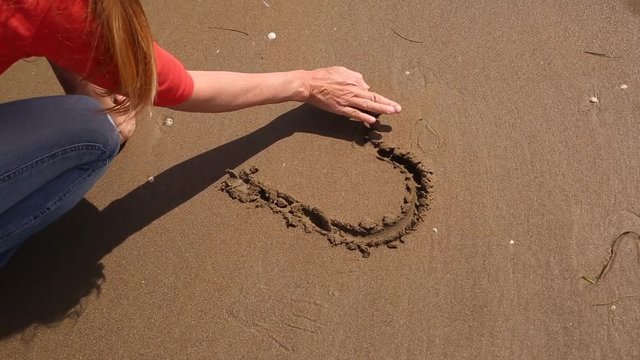 Blonde woman in love draws heart symbol with his hand on sandy ground.