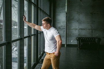 Young sexy men bodybuilder athlete,studio portrait in loft on the background of stylized wall and Black leather sofa, guy model in white T-shirt and brown trousers against background of larger window