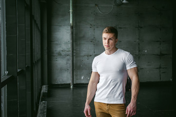 Naklejka na ściany i meble Young sexy men bodybuilder athlete,studio portrait in loft on the background of stylized wall and Black leather sofa, guy model in white T-shirt and brown trousers against background of larger window