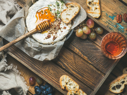 Homemade camembert cheese with honey and herbs in small pan, grapes, baguette slices, pecan nuts and glass of rose wine over rustic wooden background, top view, copy space
