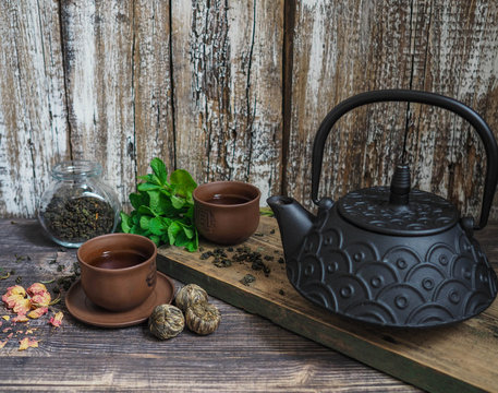 A black teapot with green tea and a cup for tea next to a sprig of mint on a wooden table
