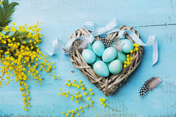 Painted Easter eggs in heart nest and mimosa flower on vintage blue background top view in flat lay style.