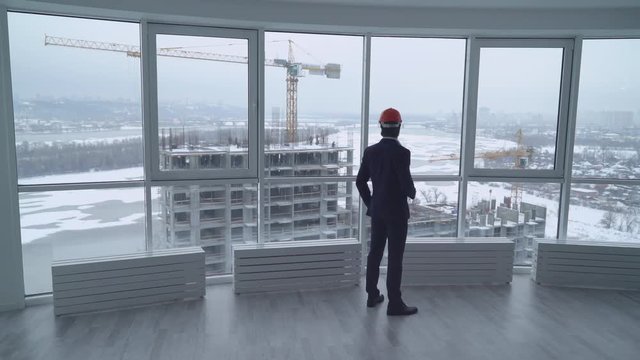 businessman in the white office looking on the street with construction site wearing in elegant suit drinking coffee oversees how workers working. rear back view man with orange hard hat in modern