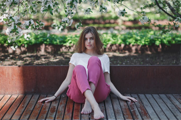 Beautiful young brunette girl sitting on a wooden bench in a blooming cherry orchard.