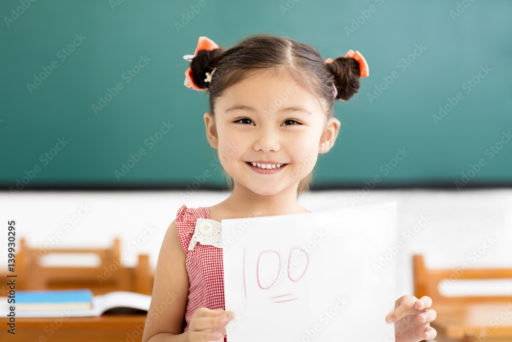 Wall mural happy little girl showing exam paper with a plus in classroom