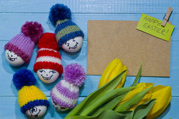 Easter eggs on wood. Emoticons in knitted hat with pom-poms. A bouquet of yellow tulips.