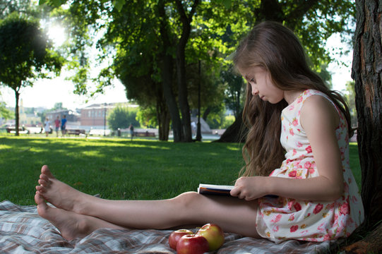 Cute little girl in a dress with flowers sitting on the grass and reading a book on a sunny summer day. On open air. training. Reading. Development