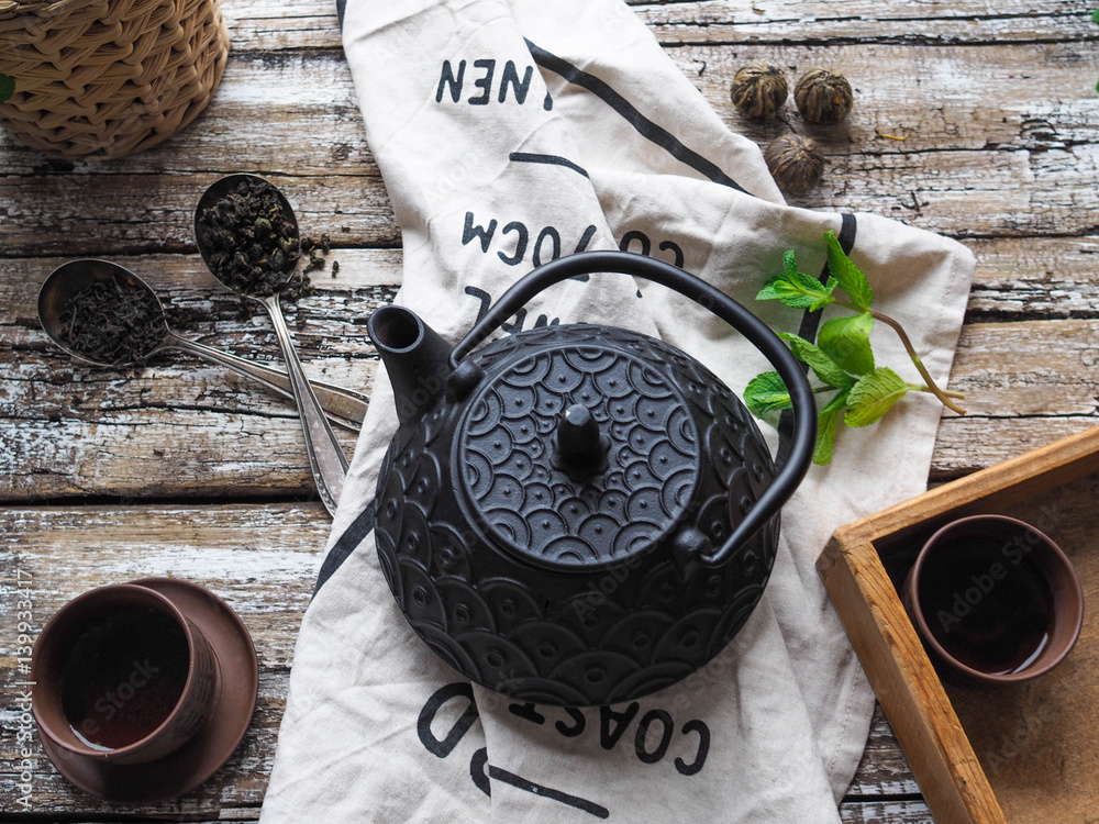 Wall mural Black teapot, two bowls with green tea, mint and spoons with scattered black and green tea on an old wooden table
