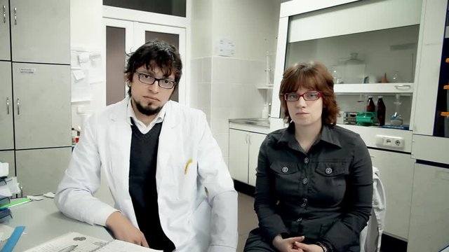 Scientific Laboratory / Research Team / Monitoring Room. Two young scientists, a man and a woman, sitting in the lab staring at camera with opposite emotions.