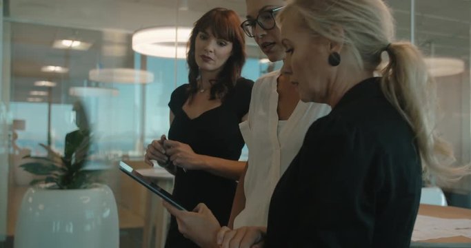 Business women standing in front of a glass wall with sticky notes and discussing. Female colleagues using digital tablet in a meeting in modern office.