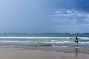 A young woman standing ankle deep in a tropical sea looking out at traditional filipino pump boats and the passing storm clouds.