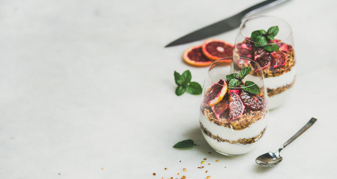 Healthy breakfast. Greek yogurt, granola, blood orange layered parfait in glasses with mint leaves over grey marble background, copy space. Clean eating, weight loss, detox, dieting food concept