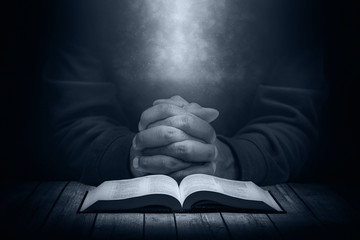 Man praying on a wooden table with an open Bible in the dark. - 139957696