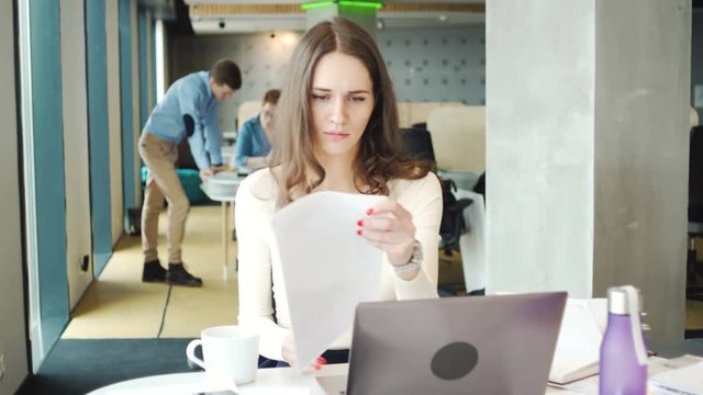 young busy hardworking woman with long dark hair sit at the desk in co working open office with laptop and business papers while solving working tasks careful and professional during sunny day