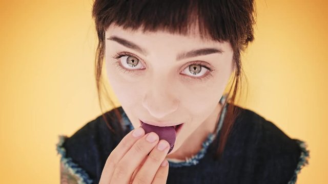 Top view close up, attractive young brunette girl with big beautiful eyes isolated on bright yellow background, looks at camera and licks violet macaron cookie