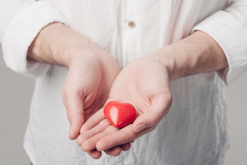 man in white shirt holds in hands red heart on gray background. 