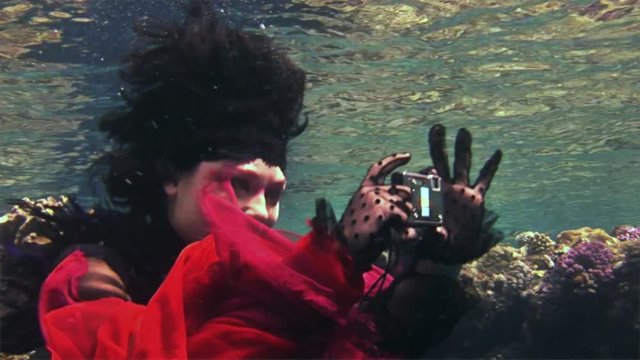 Young girl free diver in red dress photographs on camera underwater in Red Sea. Filming a movie. Actress smiling at camera. Extreme sport in marine landscape, coral reefs, ocean inhabitants.