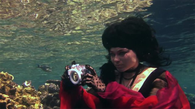 Young underwater model free diver in red dress photographs on camera in Red Sea. Filming a movie. Extreme sport in marine landscape, coral reefs, ocean inhabitants.