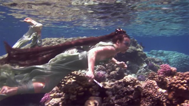Underwater model free diver in fairy costume on background of corals in Red Sea. Filming a movie. Young girl smiling at camera. Extreme sport in marine landscape, coral reefs, ocean inhabitants.