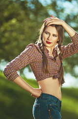 Portrait of a young beautiful woman, dressed in country style, with freckles and pigtails, standing in the meadow in a spring sunny day with tree on background