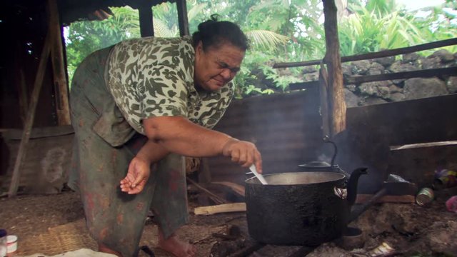 Samoan woman stirring a pot over a cooking fire in her home