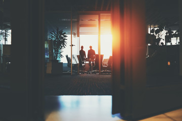 Rear view from bottom through half-closed doors of silhouettes of two young businessmen near big window in dark office interior after meeting having rest, discussing something and looking outside
