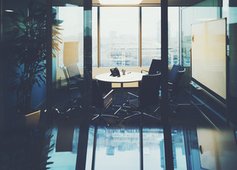 View of meeting room with round table, plant, blank white board and multiple armchairs in modern office interior with strong reflection below and huge windows with urban cityscape outside