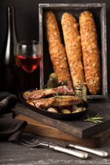 Grill pork steak on an old rustic wooden chopping board in a country kitchen with red wine and french baguette on wooden black background. Pork steak in low key