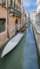 Fototapeta na wymiar Narrow Venetian canal in the historic center with row of boats moored and covered next to the typical stone buildings, Venice, Italy