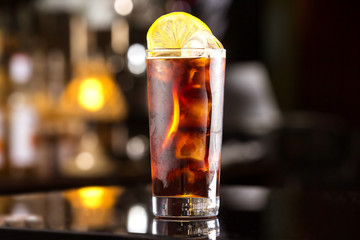 Rum cola casual cocktail with ice cubes and lemon at festive bar stand background.