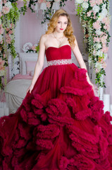 Charming woman in a beautiful lush dress cloud royal burgundy. She stands by the bed, decorated with flowers
