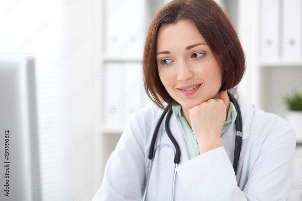 Wall mural Young brunette female doctor sitting at a desk and working on the computer at the hospital office.  Health care, insurance and help concept. Physician ready to examine patient