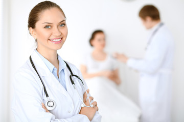 Fototapeta premium Cheerful smiling female doctor on the background with physician and his patient in the bed. High level and quality of medical service concept.