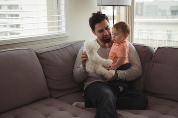 Father and baby playing with teddy bear on sofa in living room