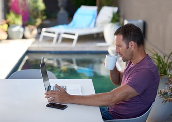 Man working on laptop computer connected on holiday vacation reading emails beside villa resort pool