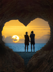 Paar Silhouette schaut am Meer vor einer Herz- förmigen Höhle in den Sonnenaufgang 