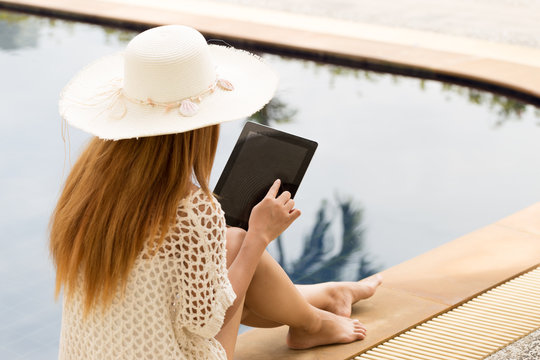 Young woman sitting by poolside in summer hat, reading on a tablet. Relaxing holiday scene blending leisure and technology lifestyle. Rear view, natural light. - Powered by Adobe