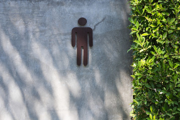 Male restroom icon mounted on a concrete wall with sunlight and leafy green hedge, symbolizing...