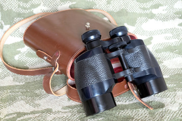Vintage military Porro prism black color binoculars with brown leather carry case with strap on camouflage background side view close up - Powered by Adobe
